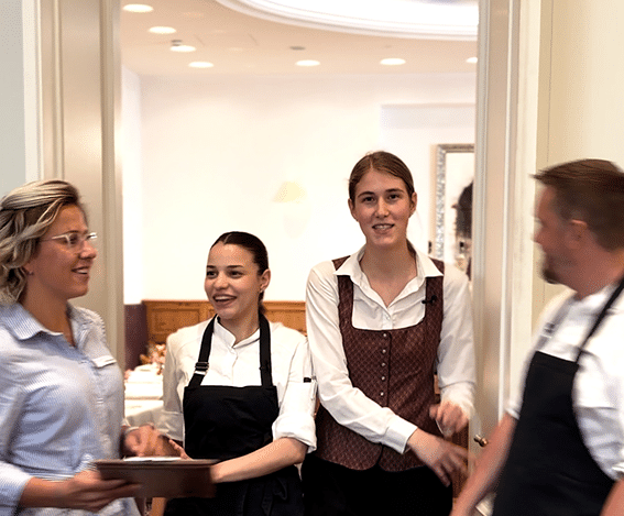 Vier Personen; Davon in der Mitte 2 junge Frauen in Arbeitskleidung von Küche und Service