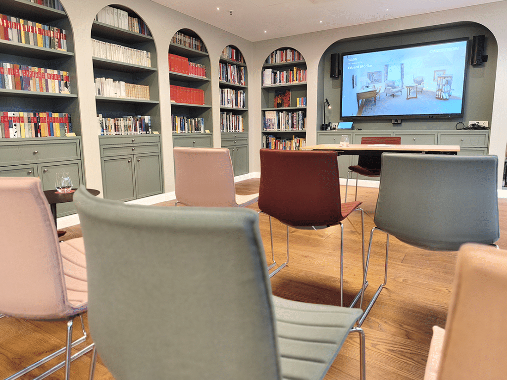 Blick druch Stuhlereihen auf einen großen Bildschirm in einem Tagungsraum. An der Seite Regale mit Büchern