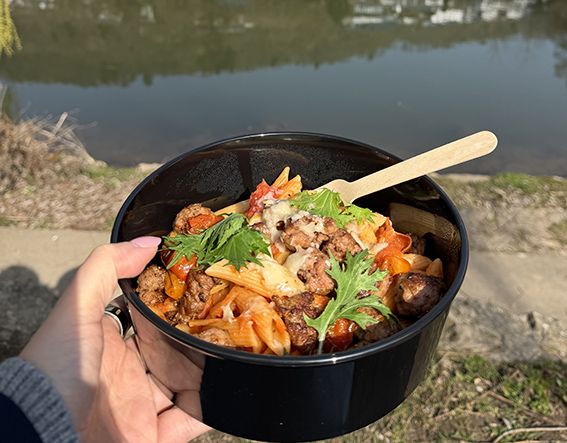 Nudelgericht in Mehrwegbehälter wird in der Hand gehalten. Im Hintergrund ist ein Fluss zu sehen.