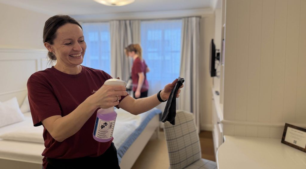 Eine Frau spürht mit einer Flasche etwas ein und lacht dabei. Im Hintergrund saugt eine andere Person
