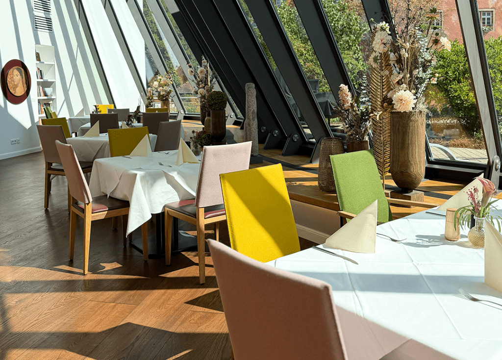 Blick in ein Restaurant mit eingedeckten Tischen. An den Stuhllehnen sind farbige Hussen angebracht. Im Hintergrund ist eine schräge Glasscheibe eines Wintergartens zu sehen.