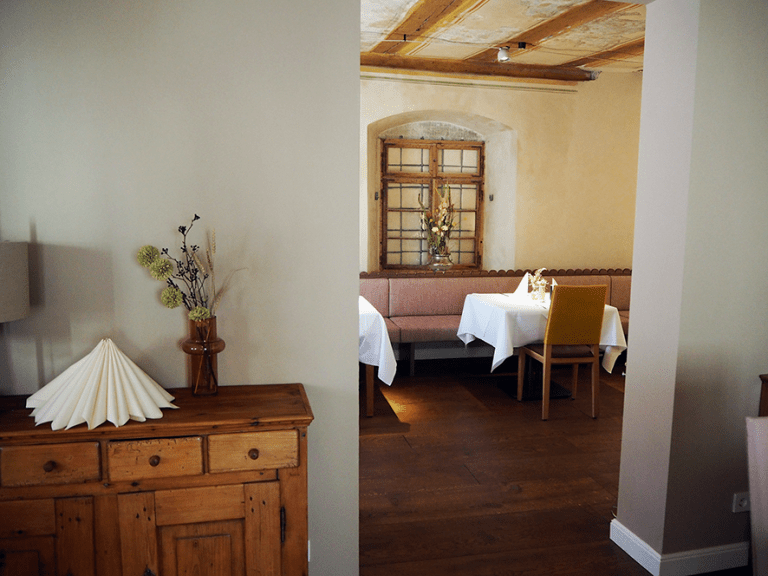 Blick durch einen Durchgang in einen Raum mit eingedeckten Tischen und Holzelementen an der Decke und im Hintergrund. Im Vordergrund eine Holzkommode.