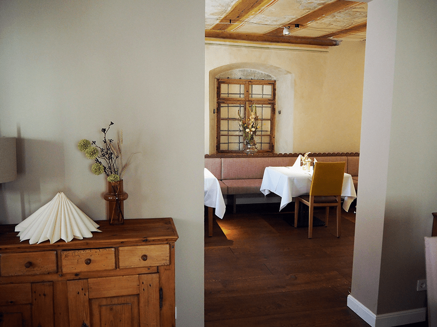 Blick durch einen Durchgang in einen Raum mit eingedeckten Tischen und Holzelementen an der Decke und im Hintergrund. Im Vordergrund eine Holzkommode.