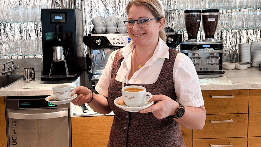 Im Vordergrund eine lächelnde Frau in Arbeitskleidung eines Cafés; in jeder Hand eine Kaffeetasse.